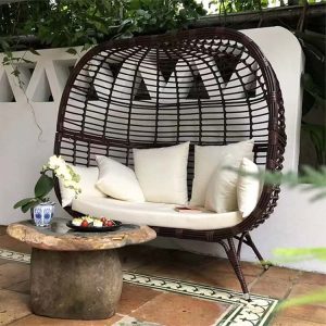 Ghế đôi lồng trứng ngoài trời màu nâu đậm, nệm trắng, đặt trong không gian xanh
