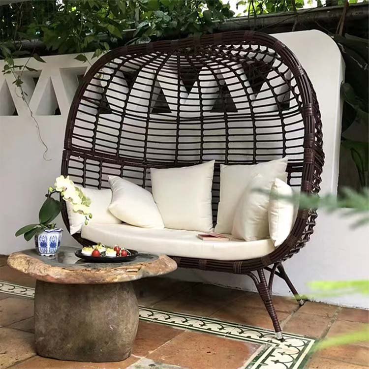 Ghế đôi lồng trứng ngoài trời màu nâu đậm, nệm trắng, đặt trong không gian xanh